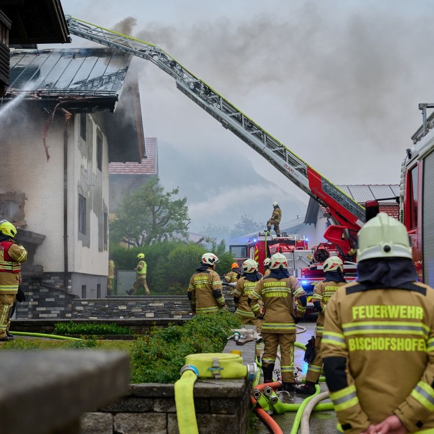 Firefighters are working on a house fire. A fire engine with a ladder is on the scene. Smoke is coming from the roof.