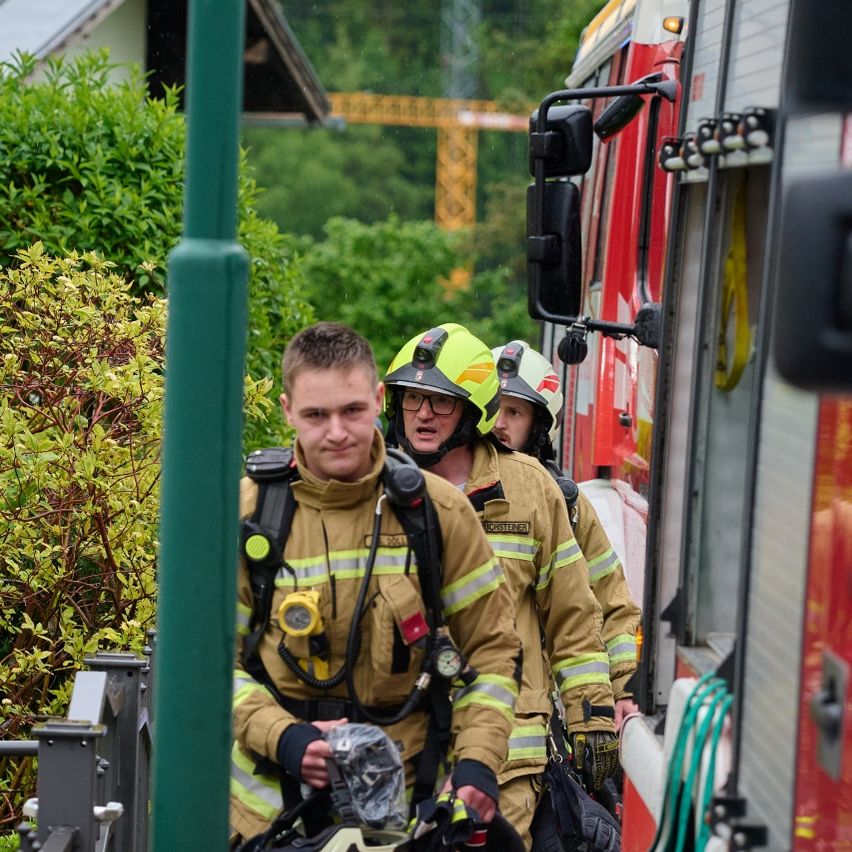 Three firefighters in protective gear walk by a fire truck near bushes. A green pole and a house are in the background.