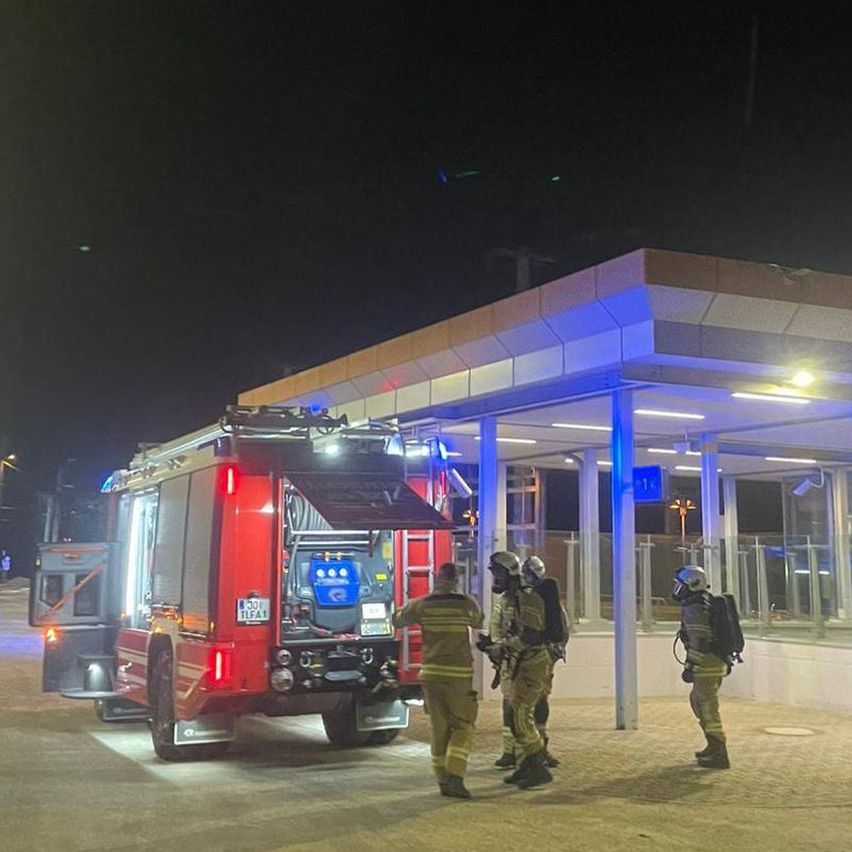 A fire truck is parked in front of a building with firefighters discussing. The building has a glass wall and a blue light.