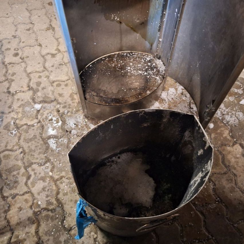 A dirty metal bucket with debris inside sits on a tiled floor, next to a partially opened metal structure with stains.