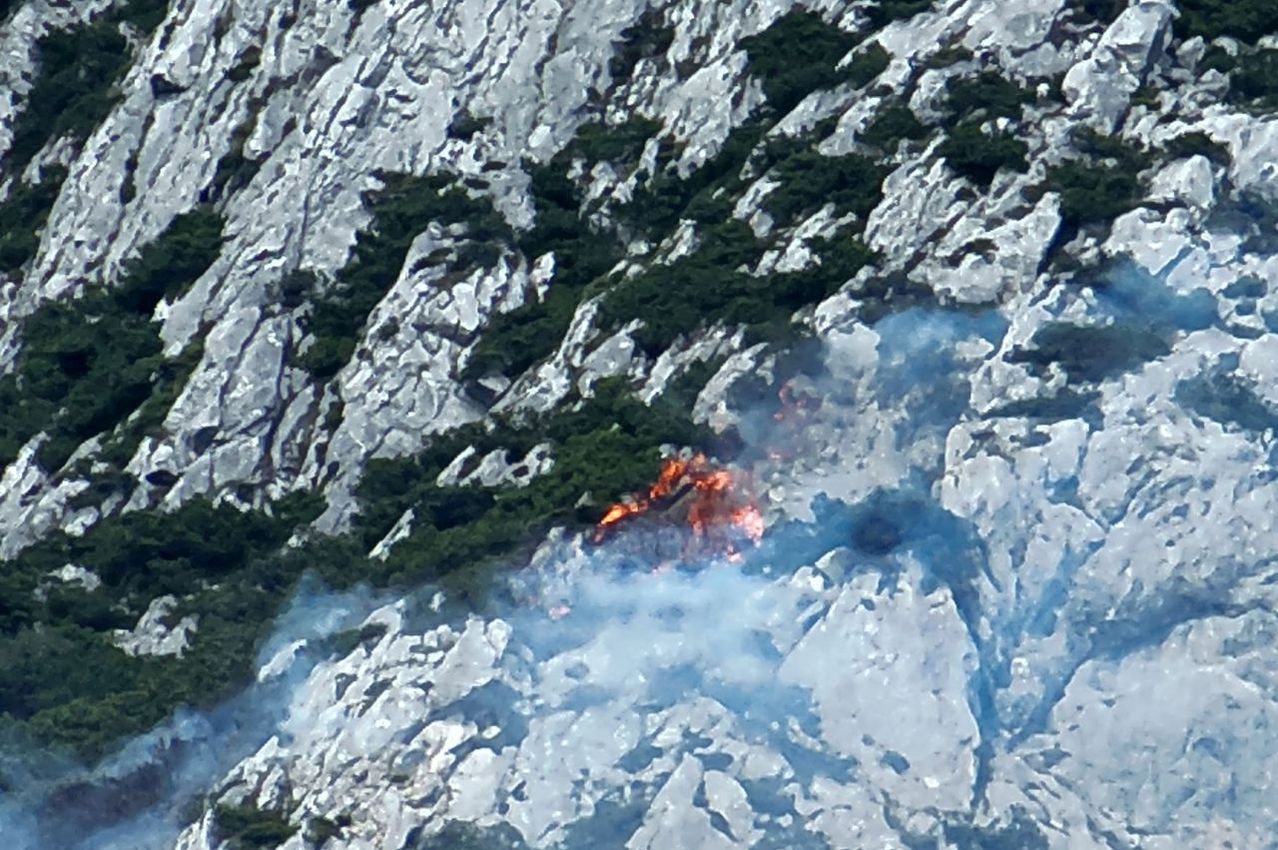 A wildfire burns through a rocky mountain area, with flames and smoke rising amidst the greenery.