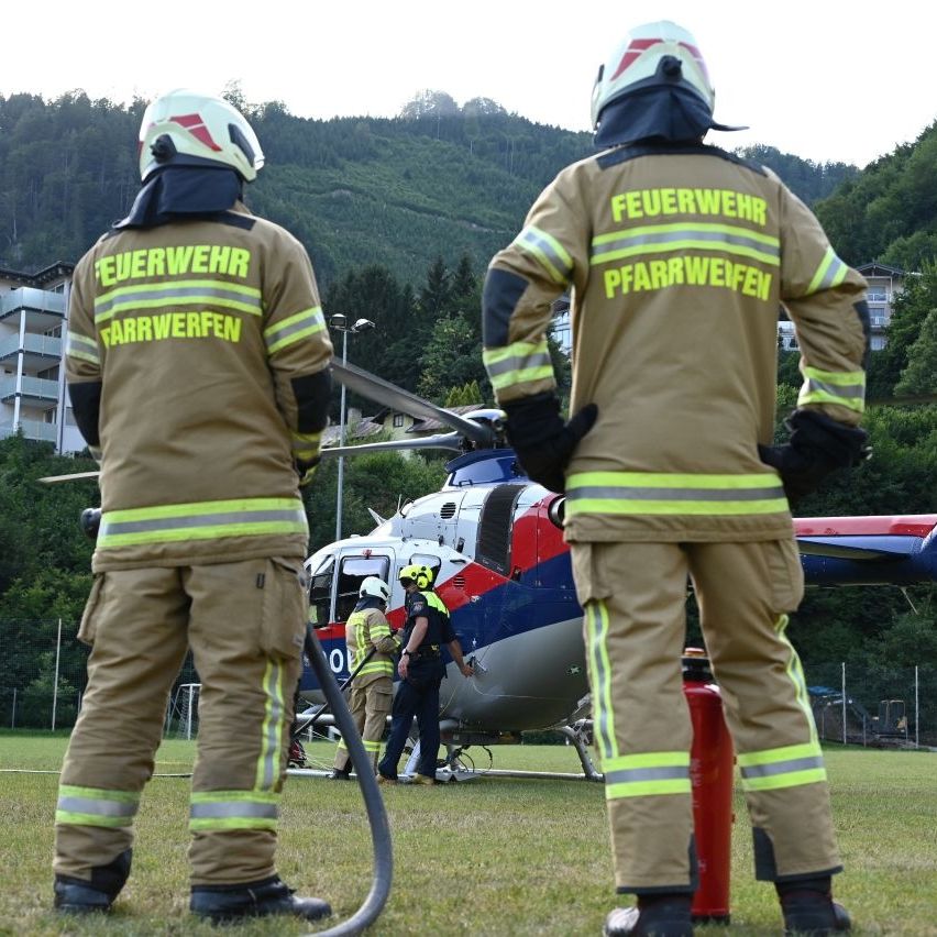 Two firefighters in full gear stand near a helicopter on a grassy field with a fire extinguisher nearby, a building and trees in the background.