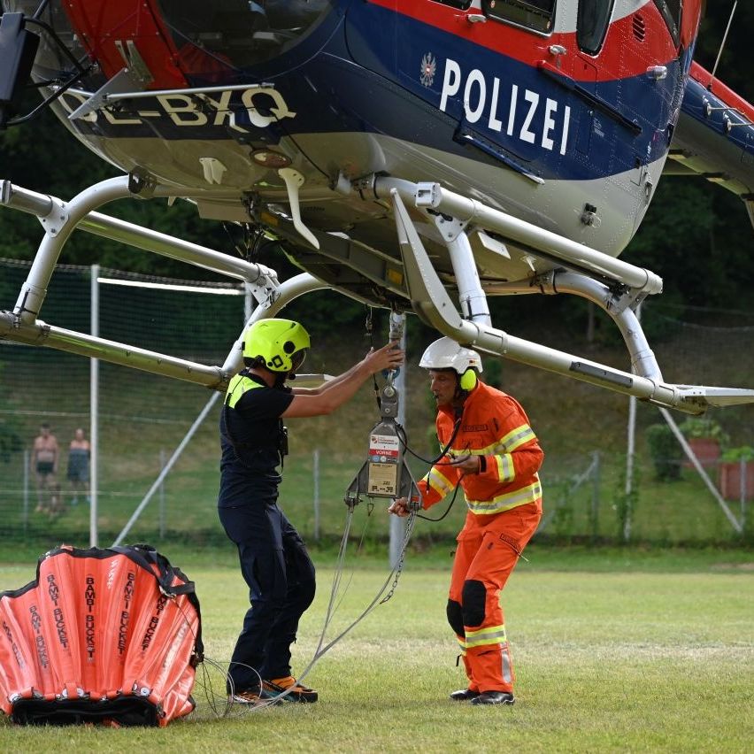 Two individuals in safety gear are securing a rope to a helicopter marked 'POLIZEI'. One is holding a chain, and the other is holding a red bag. Behind them, two people are standing near a fence.