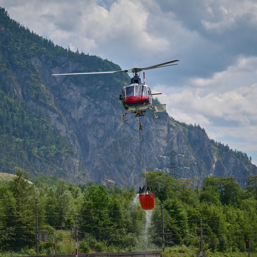 Ein Hubschrauber mit einem roten Wassereimer fliegt über ein bewaldetes Gebiet mit Bergen im Hintergrund.