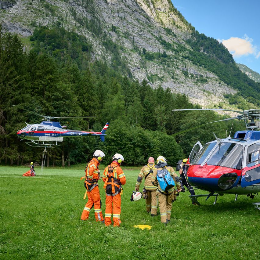 Feuerwehrleute in orange Uniformen bereiten sich darauf vor, einen Hubschrauber in einem Grasfeld mit Berghintergrund zu besteigen. Ein anderer Hubschrauber landet in der Nähe.