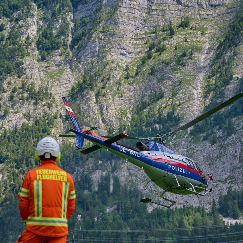 Ein Feuerwehrmann in oranger Uniform beobachtet einen Polizeihubschrauber, der über ein bergiges Gelände fliegt.