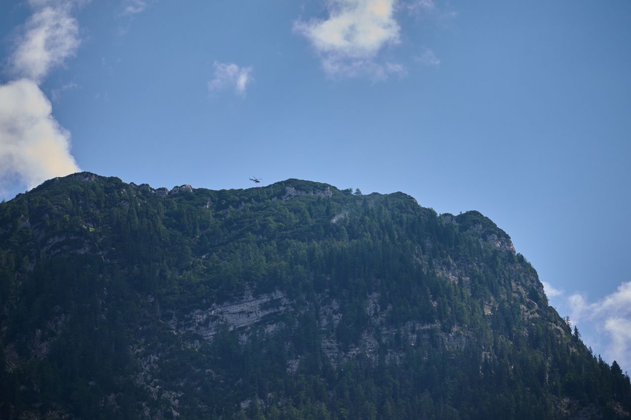 Ein Hubschrauber fliegt über einem bergigen Gelände mit dichtem Waldbestand unter einem klaren blauen Himmel.