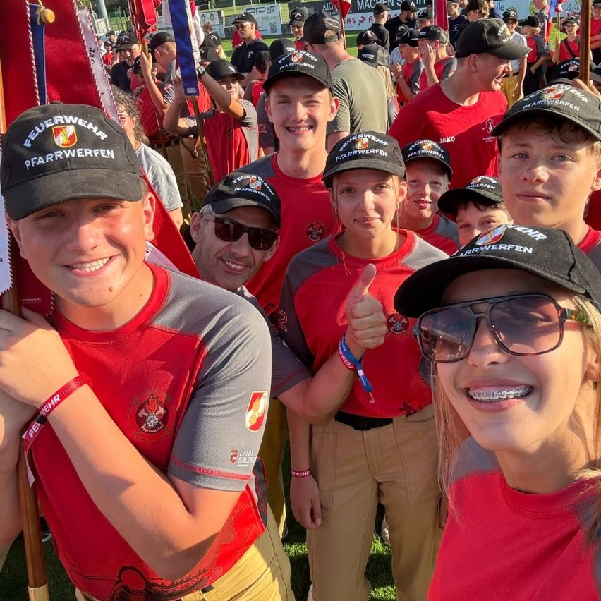 Eine Gruppe junger Menschen in roten und grauen Uniformen mit schwarzen Mützen lächelt und posiert für ein Foto. Sie halten Fahnen und stehen auf einer Rasenfläche.