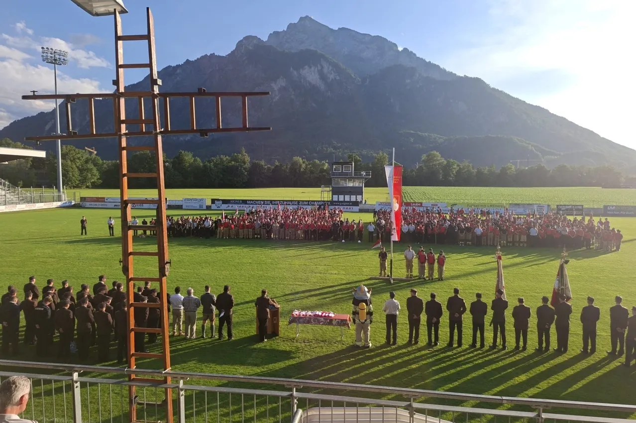 Eine große Gruppe von Menschen in roten Uniformen versammelt sich auf einem Rasenfeld mit Bergen im Hintergrund. Eine Flagge ist auf einem Pfahl gehisst, und einige Leute stehen vor einem Tisch.