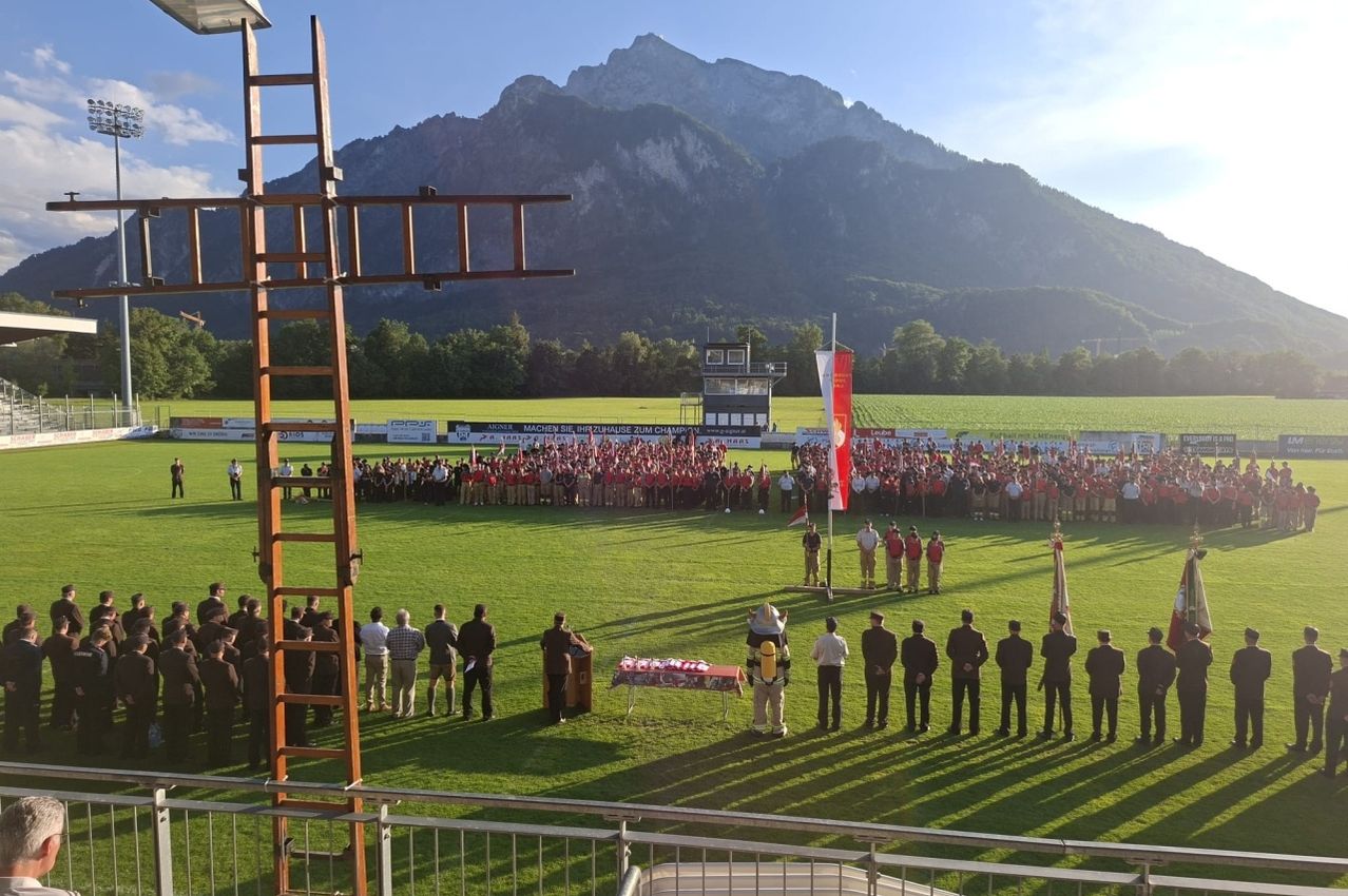 Eine große Gruppe von Menschen in roten Uniformen versammelt sich auf einem Rasenfeld mit Bergen im Hintergrund. Eine Flagge ist auf einem Pfahl gehisst, und einige Leute stehen vor einem Tisch.