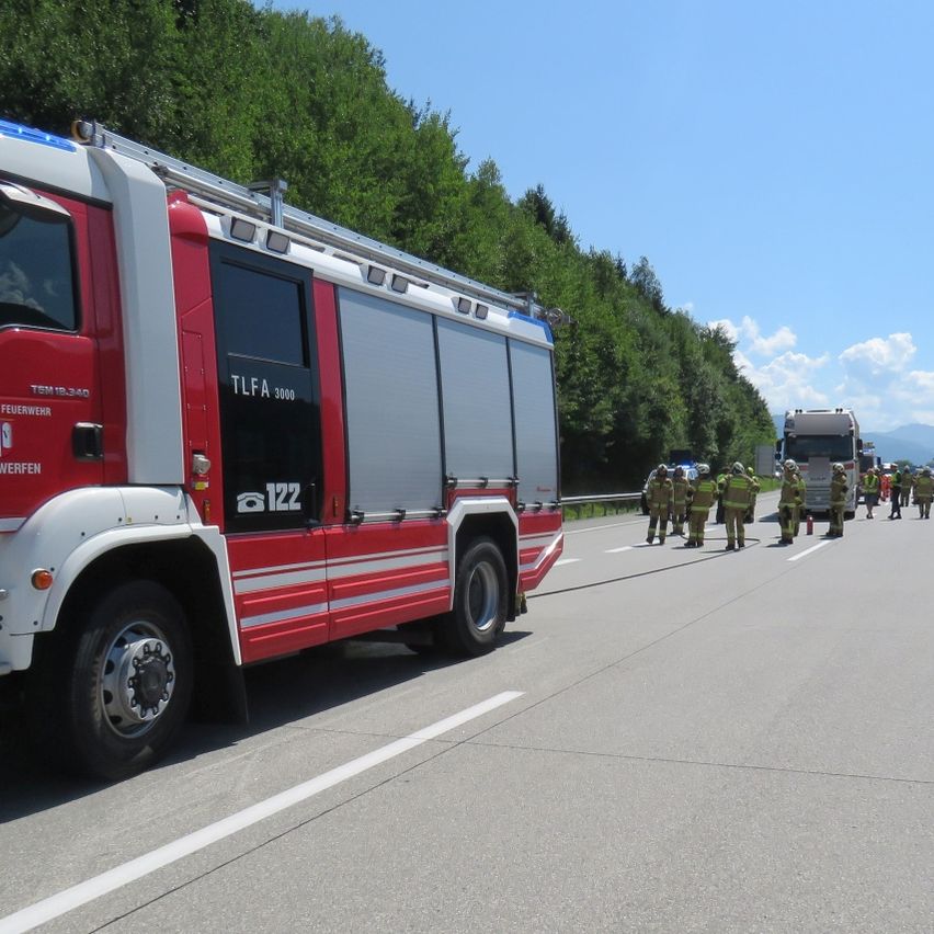 Ein roter Feuerwehrwagen mit der Aufschrift 'TLFA 3000' steht am Straßenrand. Im Hintergrund stehen Feuerwehrleute, einige halten eine Feuerwehrschläuche.