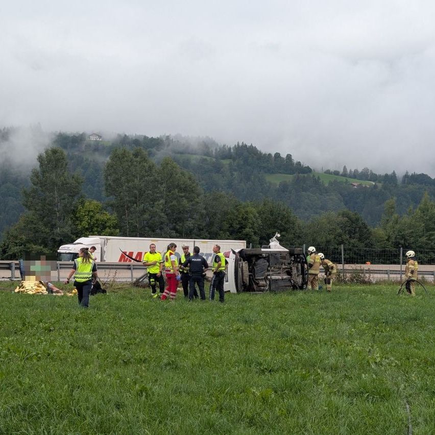 Rettungskräfte und Polizei versammeln sich um einen umgekippten Lkw auf einem Grasfeld mit einem bergigen Hintergrund.