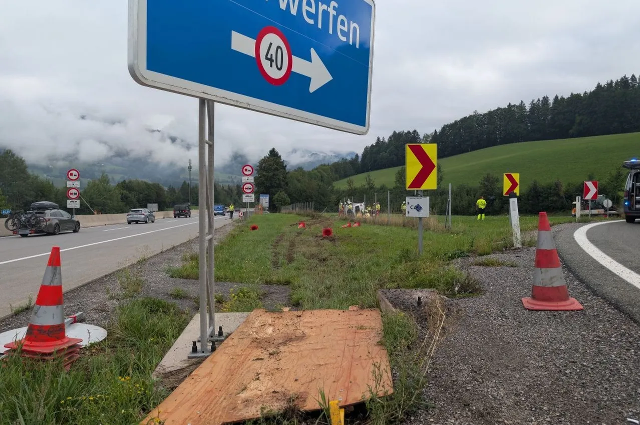 Ein Straßenschild mit einer Geschwindigkeitsbegrenzung von 40 km/h steht am Straßenrand. In der Nähe befinden sich Baukegel und Arbeiter in gelben Westen. Die Straße ist von Grün umgeben, und in der Ferne sind Berge zu sehen.