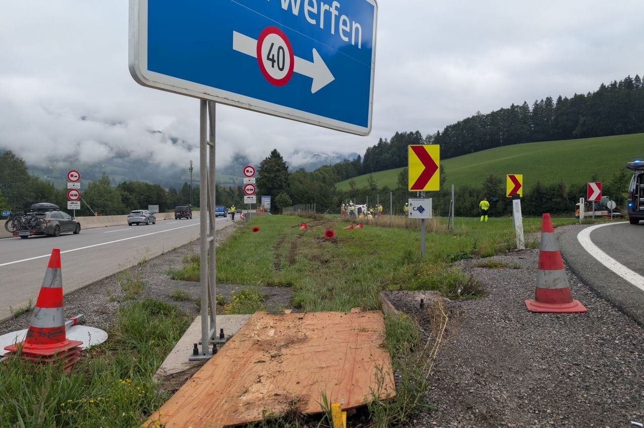 Ein Straßenschild mit einer Geschwindigkeitsbegrenzung von 40 km/h steht am Straßenrand. In der Nähe befinden sich Baukegel und Arbeiter in gelben Westen. Die Straße ist von Grün umgeben, und in der Ferne sind Berge zu sehen.