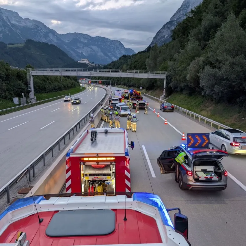 Rettungsfahrzeuge stehen am Straßenrand einer Autobahn mit Verkehrskegeln und Personen in gelben Uniformen, die sich um eine Szene kümmern. Im Hintergrund sind Berge zu sehen.