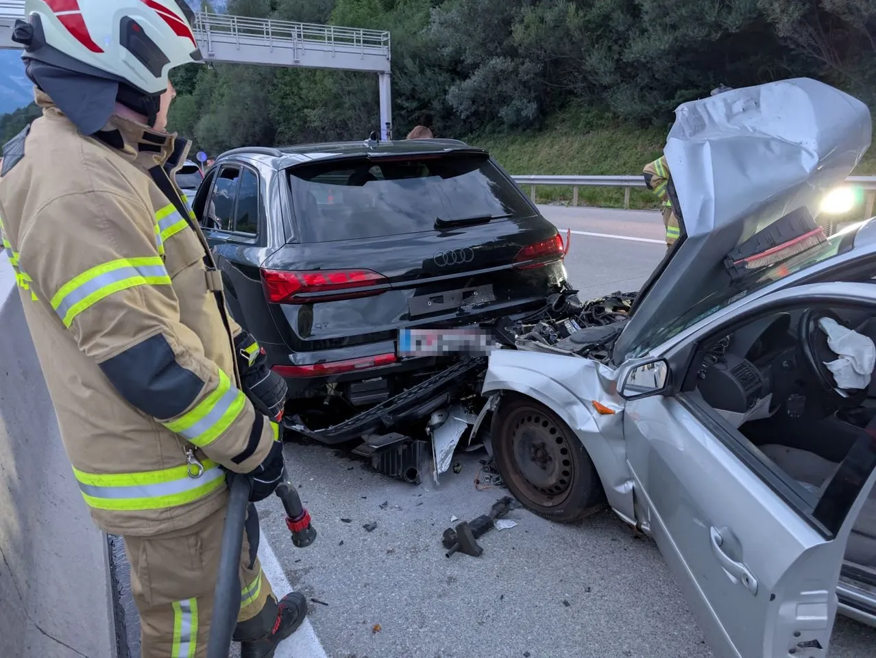 Ein Feuerwehrmann steht neben einem beschädigten schwarzen Audi SUV mit zertrümmertem Heck am Straßenrand. Ein silbernes Auto mit offenem Motorhauben liegt in der Nähe, und eine weitere Person kümmert sich darum.