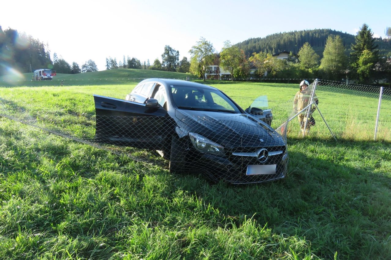 A black Mercedes-Benz car is wrecked in a grassy field with its doors open. A firefighter stands nearby, wearing a helmet and holding a tool.