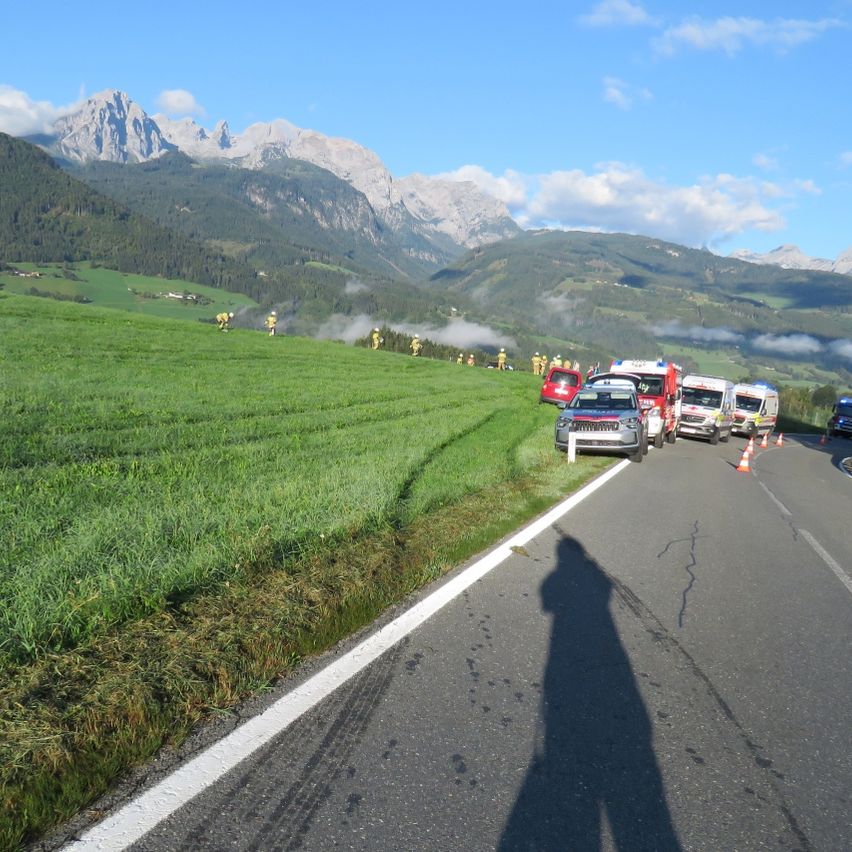 Emergency vehicles are parked on the side of a road with a shadow cast on the road. There are people in yellow uniforms in the grassy field. Mountains are in the background.