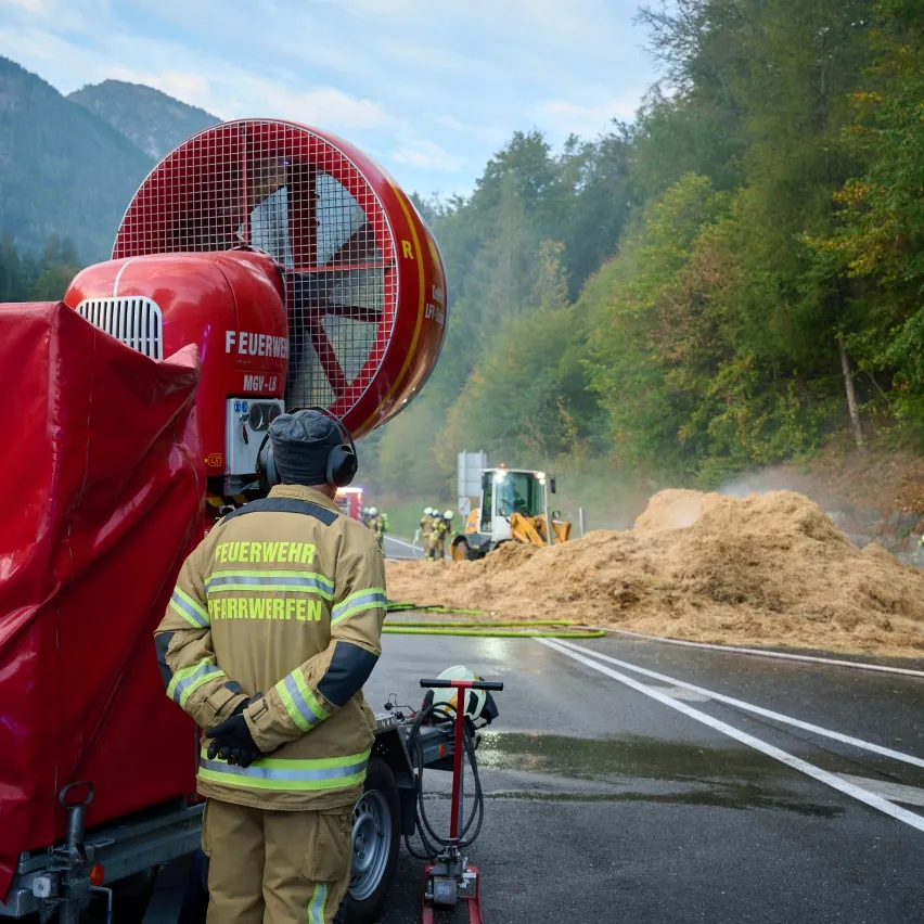 Ein Feuerwehrmann steht neben einem roten Feuerwehrauto mit einem großen Ventilator hinten, das Wasser auf einen Heuhaufen am Straßenrand sprüht.