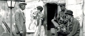 Ein Schwarz-Weiß-Bild zeigt einen Mann und eine Frau in der Nähe eines Plakats, wobei zwei Männer auf hölzernen Kisten im Hintergrund sitzen.