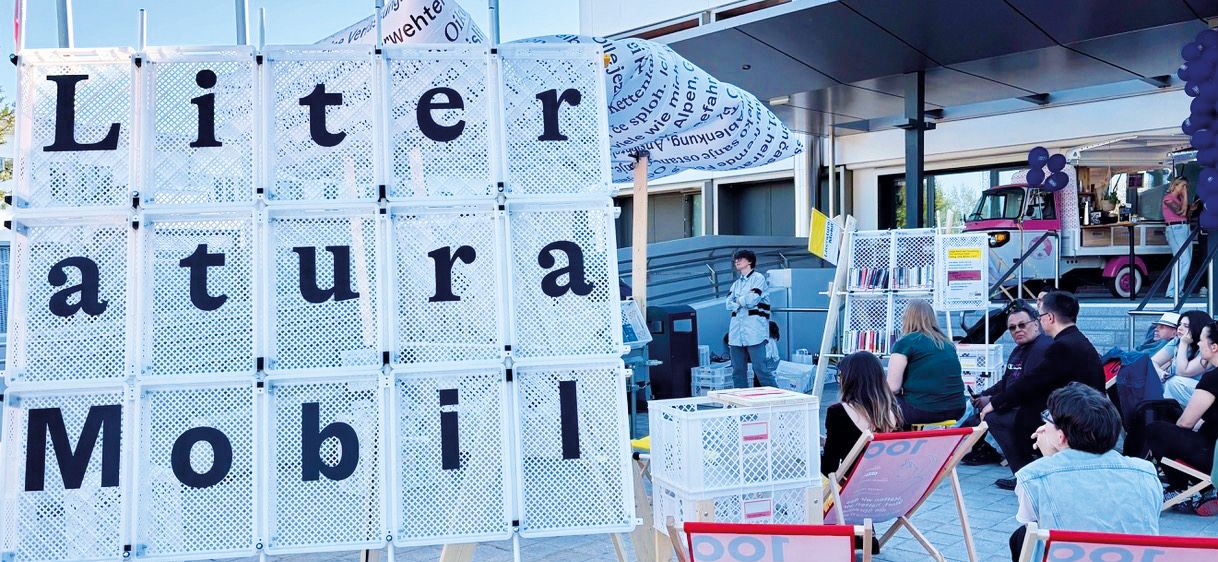 Eine Gruppe von Menschen sitzt auf Stühlen vor einem Gebäude. Ein weißes Schild mit dem Wort 'Literaturmobil' ist davor angebracht. Eine Frau steht in der Nähe des Schildes, und ein Mann hält ein Schild in der Nähe eines Bücherwagens.