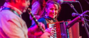 Eine Frau spielt Akkordeon und lächelt, während ein Mann neben ihr eine Klarinette auf einer Bühne mit lila Beleuchtung spielt.
