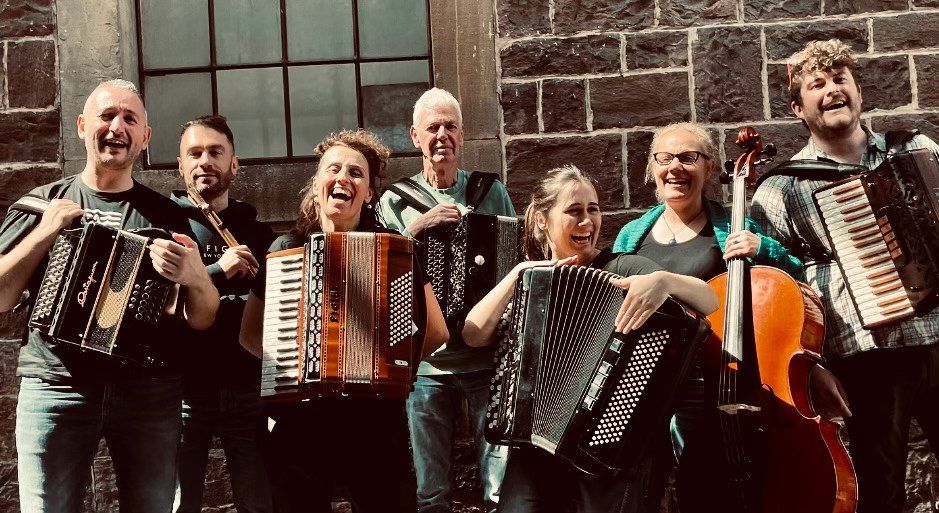 Eine Gruppe von Musikern, darunter Männer und Frauen, lächelt und posiert für ein Foto, während sie verschiedene Musikinstrumente wie Akkordeons und eine Geige vor einer Steinmauer halten.