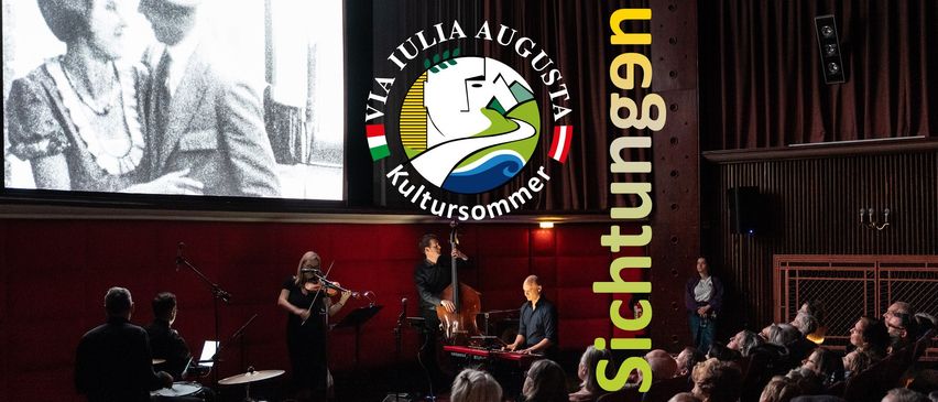 Eine Band spielt auf der Bühne mit einem großen Logo für Via Iulia Augusta Kultursommer im Hintergrund. Die Geigerin steht links, der Cellist in der Mitte und der Keyboarder rechts. Das Publikum sitzt vor ihnen.