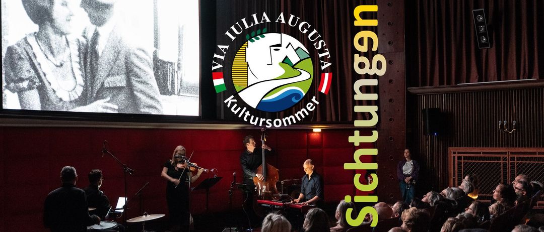 Eine Band spielt auf der Bühne mit einem großen Logo für Via Iulia Augusta Kultursommer im Hintergrund. Die Geigerin steht links, der Cellist in der Mitte und der Keyboarder rechts. Das Publikum sitzt vor ihnen.