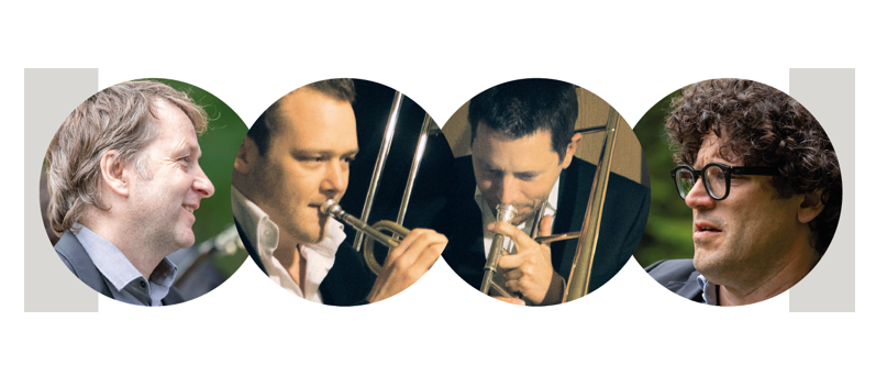 Vier Männer in Anzügen, jeder spielt ein anderes Blechblasinstrument. Von links nach rechts spielen sie Trompete, Posaune, Trompete und Posaune.