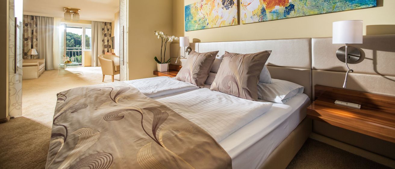 Ein Schlafzimmer mit einem Doppelbett, zwei großen Gemälden, einem Stuhl und einem kleinen Tisch mit einer Lampe. Das Zimmer hat einen Balkon mit Blick auf die Bäume.