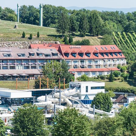Ein Hotelkomplex namens Leitner mit rotem Dach ist von Bäumen umgeben. Vor dem Hotel befindet sich eine Wasserrutsche.