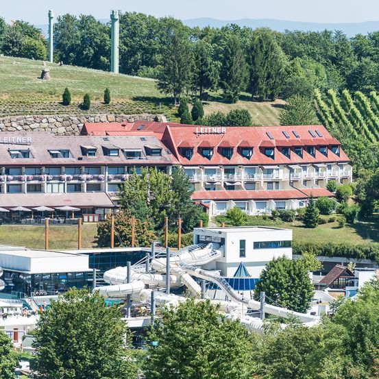 Ein Hotelkomplex namens Leitner mit rotem Dach ist von Bäumen umgeben. Vor dem Hotel befindet sich eine Wasserrutsche.