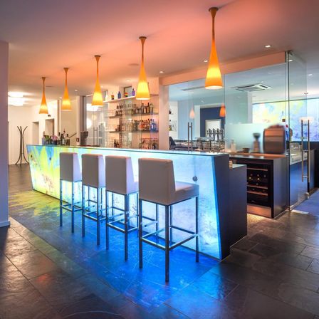 Eine moderne Bar mit vier weißen Stühlen, einer farbenfrohen Rückwand und gelben Pendelleuchten. Hinter der Bar sind Regale mit Flaschen zu sehen.