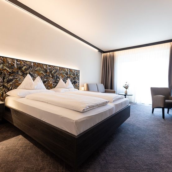 Ein Hotelzimmer mit zwei Betten, weißen Laken, dekorativen Kissen, einem Nachttisch, einem Stuhl, einem Schreibtisch, einer Vase mit Blumen und einem verhangenen Fenster.