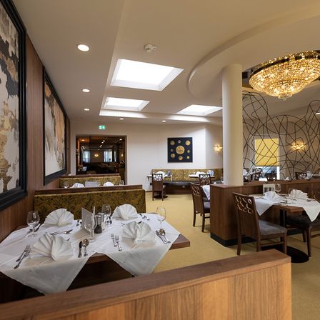 Ein leeres Restaurant mit Holzstühlen und Tischen, alles für das Essen vorbereitet. Eine große Kristallleuchter hängt von der Decke. Die Wände sind mit großen Gemälden verziert.