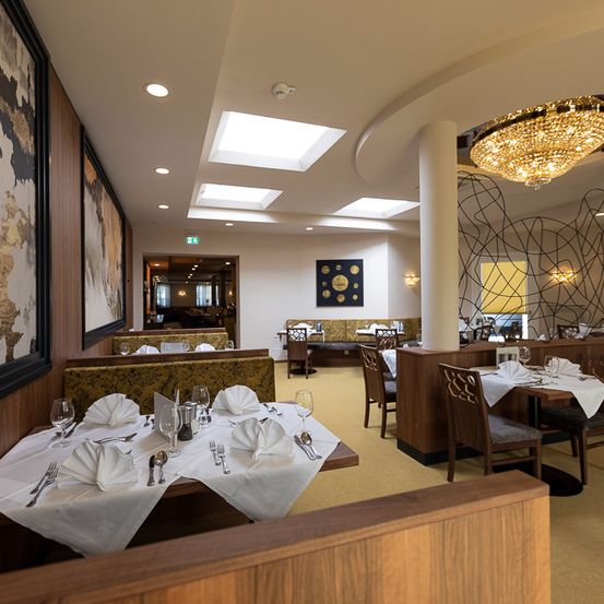 Ein leeres Restaurant mit Holzstühlen und Tischen, alles für das Essen vorbereitet. Eine große Kristallleuchter hängt von der Decke. Die Wände sind mit großen Gemälden verziert.