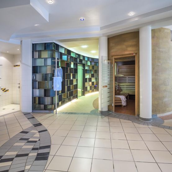 Das Innere eines modernen Spas umfasst eine große, farbenfrohe Mosaikwand, eine verglaste Sauna und einen Handtuchständer. Der Boden ist mit weißen Fliesen bedeckt.
