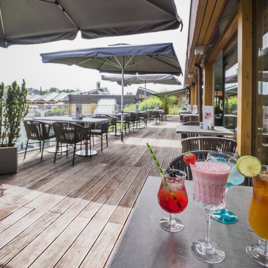 Restaurant im Freien mit Tischen, Stühlen und Sonnenschirmen. Drei Gläser bunter Cocktails mit Strohhalmen auf einem Tisch.