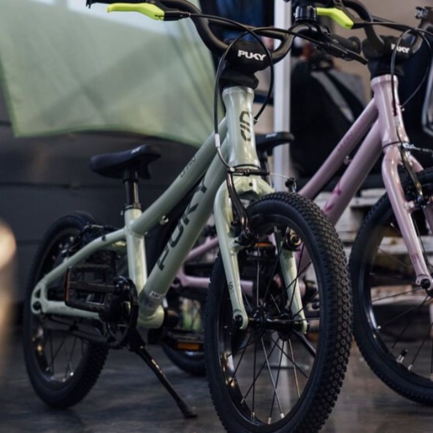 Zwei Fahrräder, eines grau und eines rosa, stehen in einem Raum. Das graue Fahrrad hat 'PUKY' auf seinen Lenkergriffen und einen gelben Griff. Das rosa Fahrrad hat schwarze Lenker.
