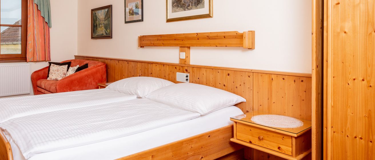 Ein gemütliches Schlafzimmer mit einem Doppelbett und Kissen, Holzwänden und einem Nachttisch. Drei gerahmte Bilder schmücken die Wand. Ein orangefarbener Sessel mit Kissen steht neben dem Fenster.