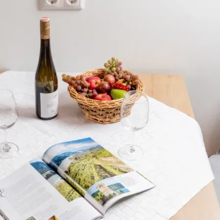 Ein Tisch mit einer Weinflasche, zwei Weingläsern, einem Obstkorb und einer offenen Zeitschrift mit einer landschaftlichen Titelseite.