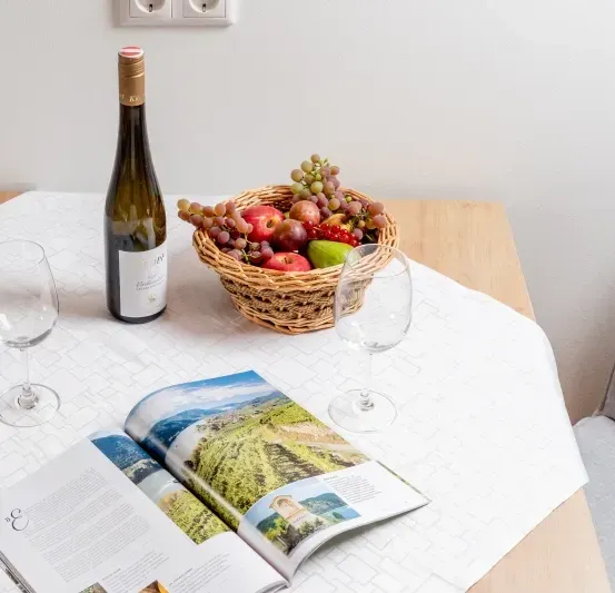 Ein Tisch mit einer Weinflasche, zwei Weingläsern, einem Obstkorb und einer offenen Zeitschrift mit einer landschaftlichen Titelseite.