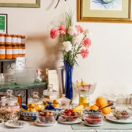 Ein Frühstückstisch mit verschiedenen Lebensmitteln. Schüsseln mit Früchten und Beeren, ein Glas mit Müsli und eine Vase mit Blumen. Dahinter sind zwei Bilderrahmen an der Wand montiert.