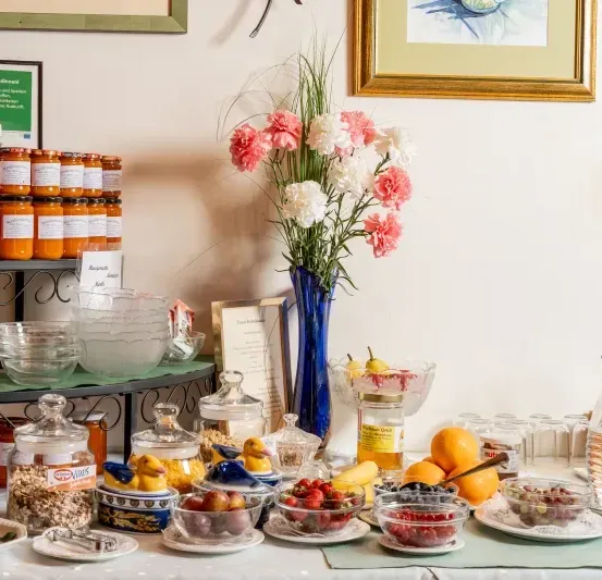 Ein Frühstückstisch mit verschiedenen Lebensmitteln. Schüsseln mit Früchten und Beeren, ein Glas mit Müsli und eine Vase mit Blumen. Dahinter sind zwei Bilderrahmen an der Wand montiert.