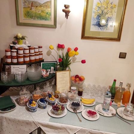 Ein Tisch ist mit einer Auswahl an Obst, Cerealien und Gewürzen gedeckt. Ein Menü ist ausgestellt. Zwei gerahmte Bilder hängen an der Wand, eines davon zeigt Blumen.