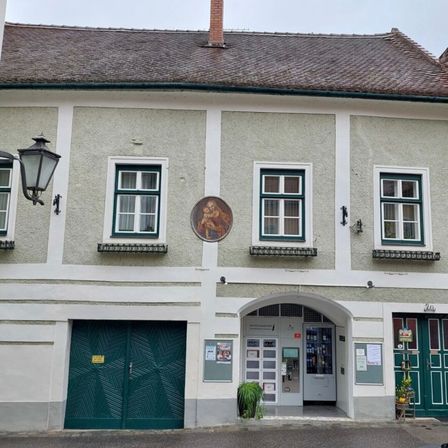 Ein Gebäude mit einem Ziegeldach und grünen Türen hat Fenster mit grünen Rahmen und ein kreisförmiges Porträt über dem Eingang.