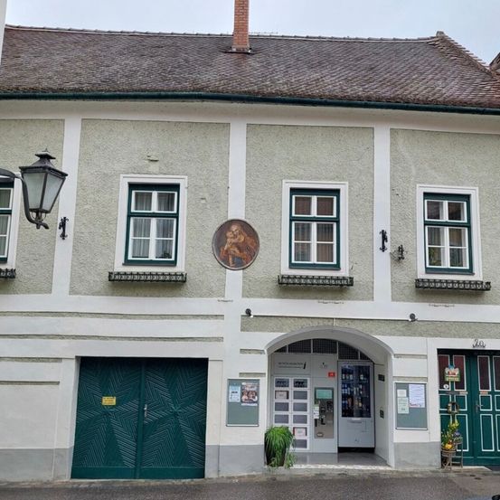 Ein Gebäude mit einem Ziegeldach und grünen Türen hat Fenster mit grünen Rahmen und ein kreisförmiges Porträt über dem Eingang.