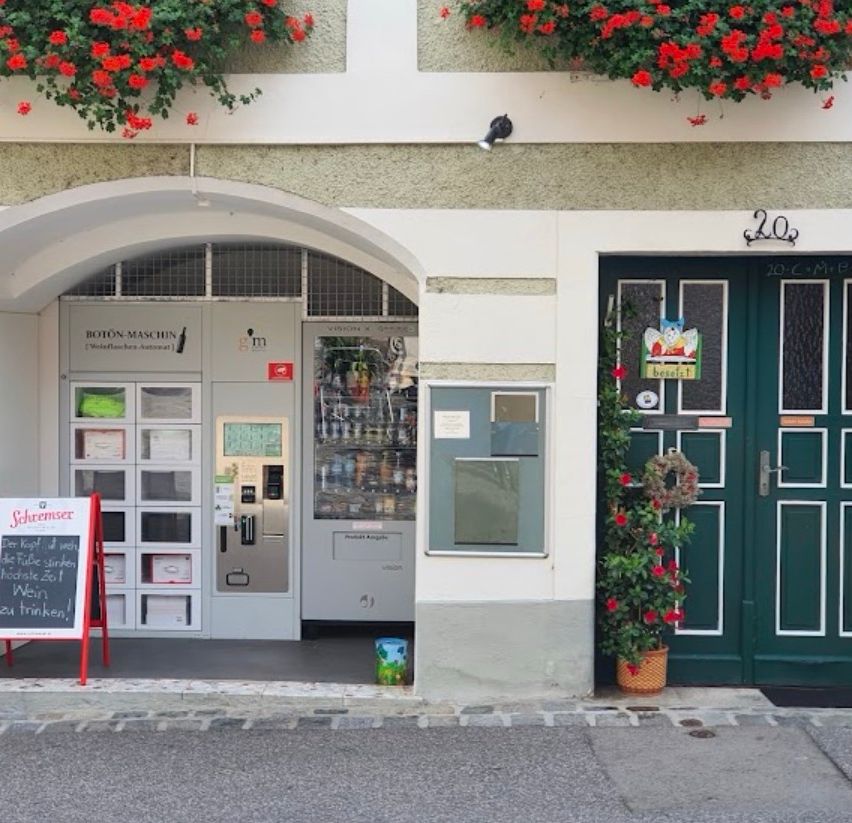 Der Eingang zu einem Geschäft mit einem rotblumigen Bogen und einem Verkaufsautomaten für Weinflaschen. Neben dem Geschäft stehen zwei grüne Türen.