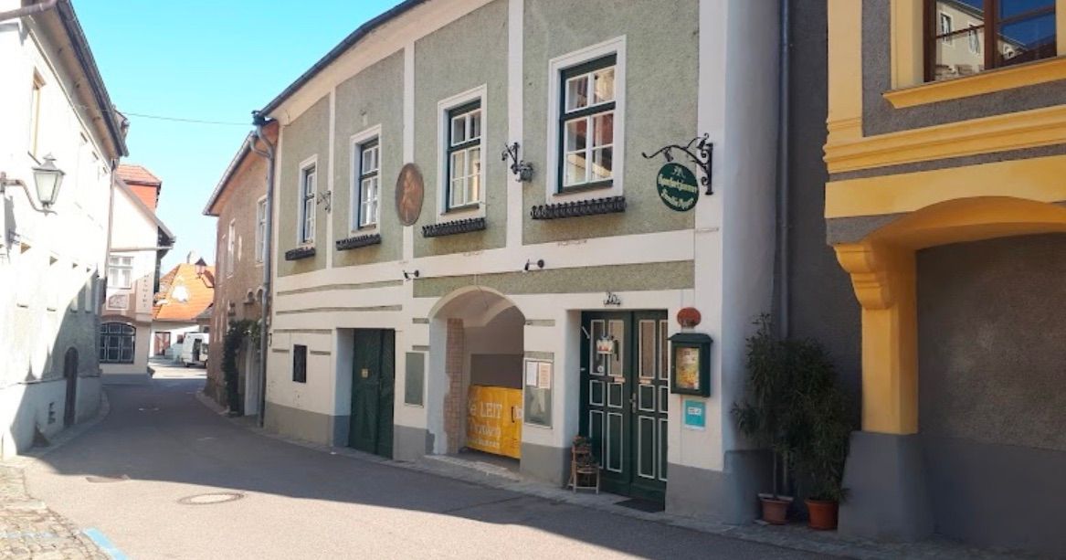 Ein Gebäude mit mehreren Fenstern und Türen. Das Gebäude ist grün und weiß gestrichen. Es hat ein Schild über der Eingangstür. Eine Pflanze ist vor dem Gebäude aufgestellt.